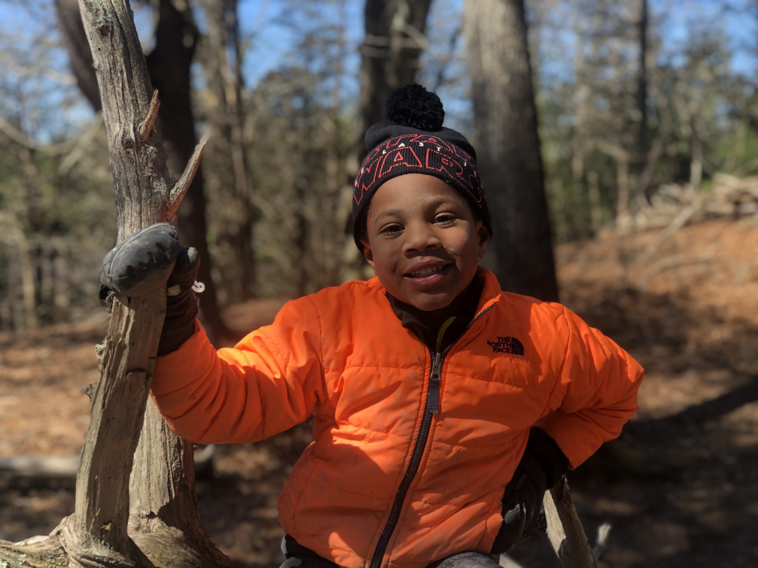 A happy child in the forest wearing an orange coat and black hat, attending nature school.